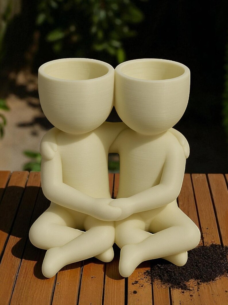 May include: A cream-coloured ceramic planter shaped like two figures embracing. Each figure has a rounded head with a hollowed-out top for planting. The figures are seated with their legs crossed and hands clasped, creating a unique decorative piece.