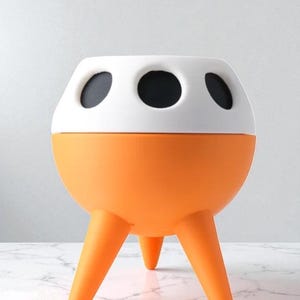 May include: A modern, orange and white decorative planter with three legs. The top section is white with three black circular openings. The lower section is orange and spherical. The planter is designed to hold plants.