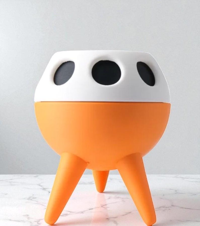 May include: A modern, orange and white decorative planter with three legs. The top section is white with three black circular openings. The lower section is orange and spherical. The planter is designed to hold plants.