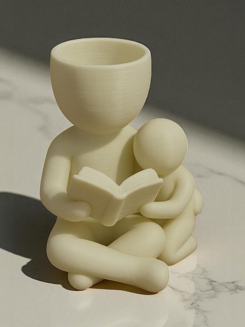 May include: A cream-colored figurine depicting a person reading a book to a smaller figure. The figures are stylized with smooth, rounded forms. The larger figure has a bowl-shaped head, suggesting a planter or container. The book is open, and the smaller figure is nestled close.