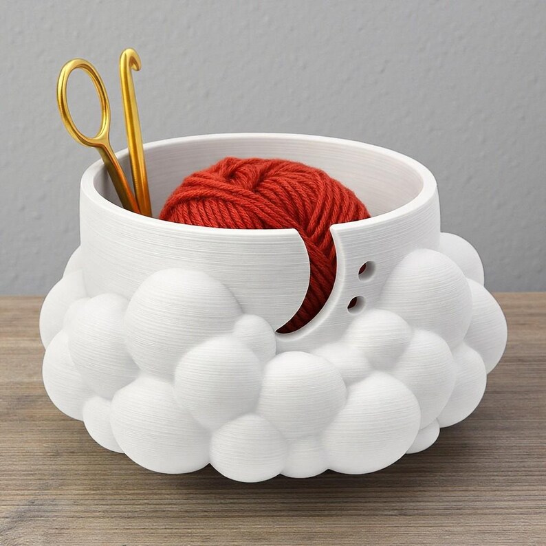 K&ouml;nnte beinhalten: Eine wei&szlig;e Garnschale mit wolkenartigem Design, die einen roten Wollkn&auml;uel enth&auml;lt. Goldfarbene Schere und H&auml;kelnadel liegen darin. Die Schale hat eine gebogene &Ouml;ffnung und zwei kleine L&ouml;cher. Ideal f&uuml;r Strick- und H&auml;kelfans.