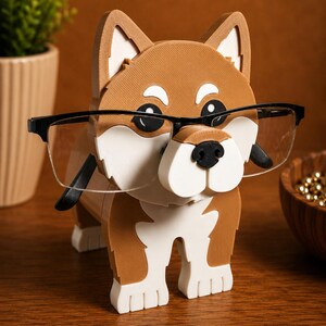 May include: A brown and white dog-shaped eyeglasses holder with black-framed glasses. The holder has a black nose and is made of wood. The dog is standing on four legs. A small plant in a beige pot is in the background.