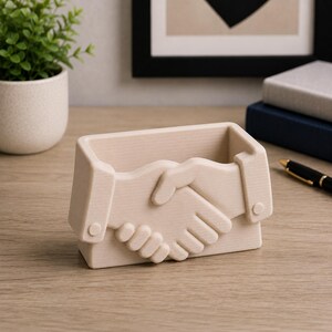 May include: Beige desk organizer shaped like two hands shaking. The organizer has a rectangular base and a hollow interior for storing pens, pencils, or other office supplies. The design features detailed relief of clasped hands.
