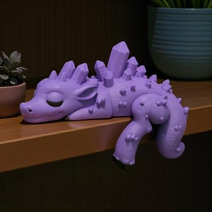 May include: A lavender-colored dragon figurine with crystal-like spikes along its back and head. The dragon is lying down with its eyes closed, and its tail and legs are curled. The figurine is resting on a wooden shelf.