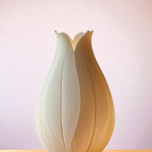 May include: A white, tulip-shaped vase with a textured surface. The vase has a wide, rounded base that tapers upwards, with petal-like edges at the top. The vase is set against a soft pink background and a yellow surface.