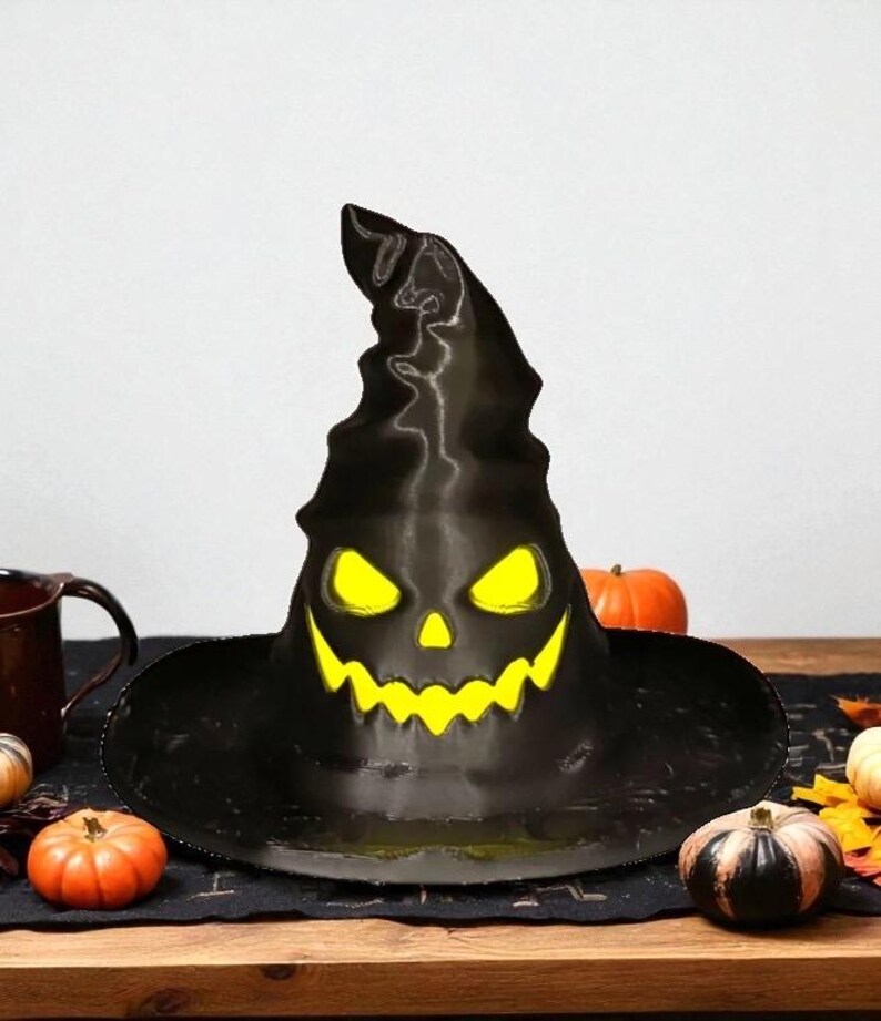 May include: A black Halloween witch's hat with a carved jack-o'-lantern face, featuring glowing yellow eyes and a toothy grin. The hat is displayed on a wooden table with pumpkins and autumn leaves, creating a festive seasonal display.