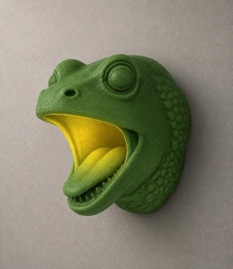 Op de afbeelding: Een groene kikkerhoofdsculptuur met een open mond en gele binnenkant, gemonteerd op een lichtgrijze muur. De huid van de kikker heeft een textuur. De sculptuur is een decoratief item.