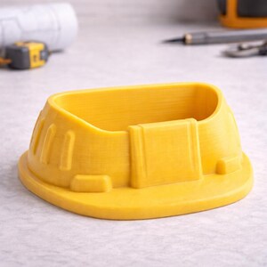 May include: A bright yellow, 3D-printed desk organizer shaped like a construction hard hat. The organizer has a rounded base and a rectangular opening for storing pens, pencils, or small items. The hard hat design includes molded details.