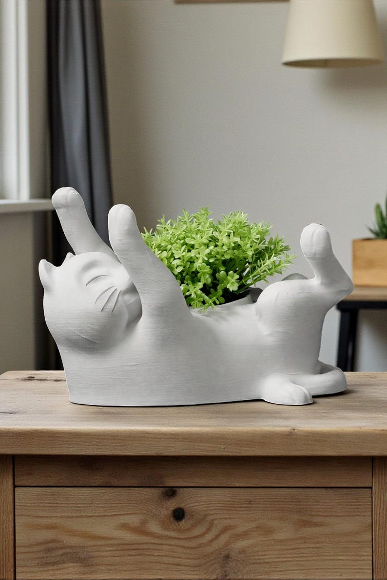May include: A white ceramic cat-shaped planter, lying on its back with paws raised. The planter holds a vibrant green plant. The cat has a smiling expression and is placed on a wooden surface.