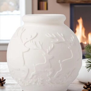 May include: A white ceramic vase with a round body and a slightly flared top. The vase features a raised relief design of reindeer and foliage. The background includes a fireplace and a window, suggesting a winter or holiday theme.