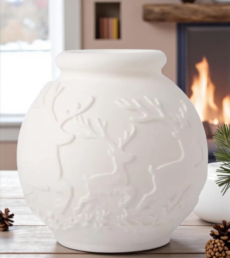 May include: A white ceramic vase with a round body and a slightly flared top. The vase features a raised relief design of reindeer and foliage. The background includes a fireplace and a window, suggesting a winter or holiday theme.