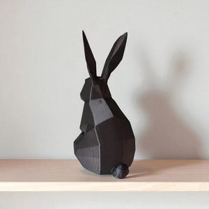 May include: A black geometric rabbit sculpture with long, pointed ears and a small, round tail. The modern art piece is displayed on a light-colored wooden shelf against a neutral wall. The rabbit's form is created from angular planes.