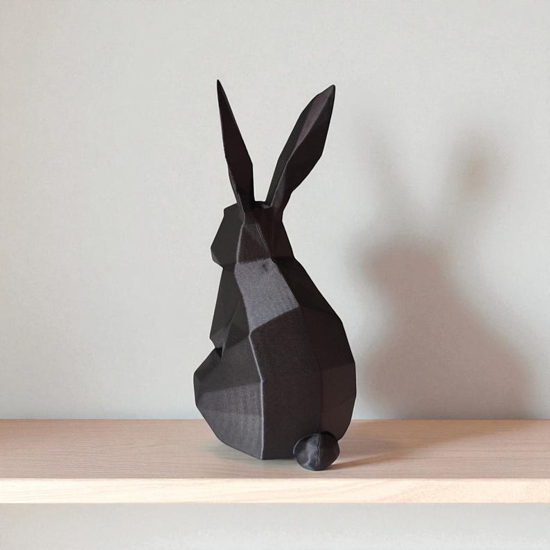 May include: A black geometric rabbit sculpture with long, pointed ears and a small, round tail. The modern art piece is displayed on a light-colored wooden shelf against a neutral wall. The rabbit's form is created from angular planes.