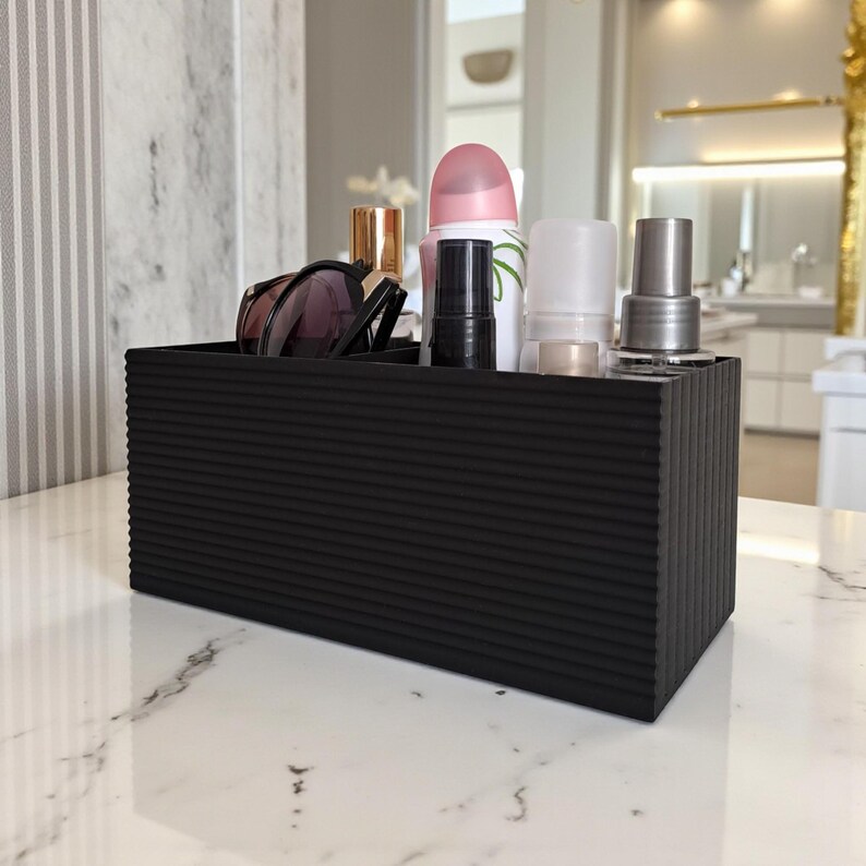 May include: A black rectangular organizer with a ribbed texture, holding various beauty products. The organizer sits on a white marble surface and contains items like sunglasses, perfume, and spray bottles. The background includes a mirror and a striped wall.