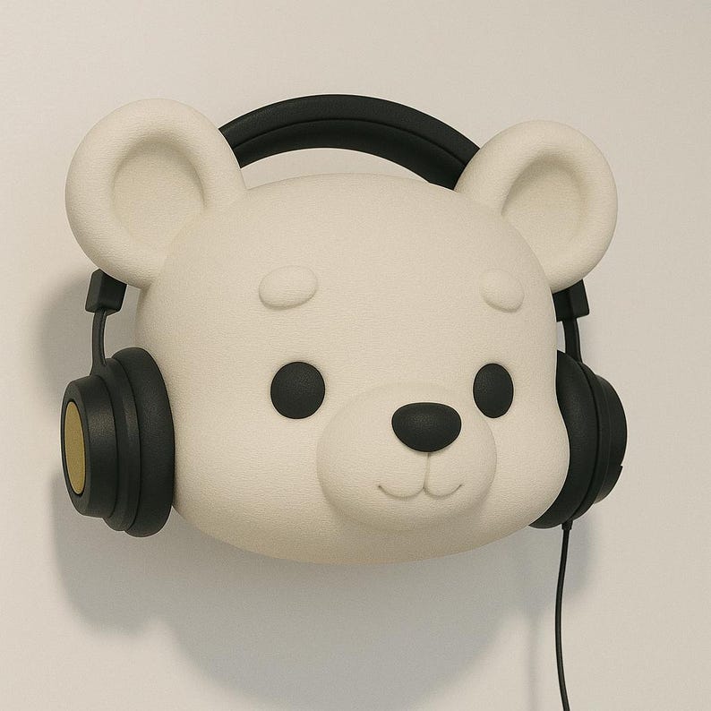 May include: A white bear-shaped wall decor with black headphones. The bear has black eyes and a black nose. The headphones have black earcups with gold accents and a black headband. The decor is mounted on a white wall.