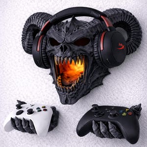 Puede incluir: Un conjunto de accesorios de juego montados en la pared con una calavera demon&iacute;aca con luz naranja brillante, que sostiene auriculares negros con detalles en rojo. Incluye dos mandos de juego, uno blanco y otro negro. El texto dice: RGB Puck Light and Remote Included!
