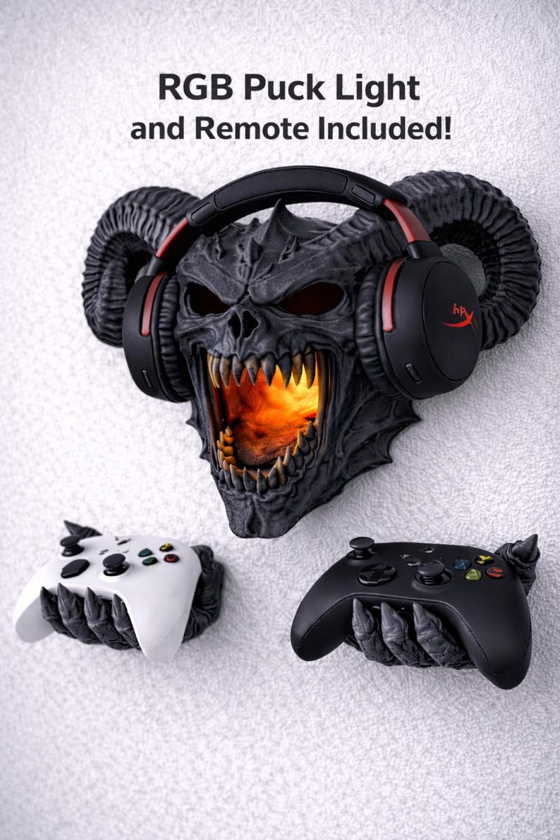 Puede incluir: Un conjunto de accesorios de juego montados en la pared con una calavera demon&iacute;aca con luz naranja brillante, que sostiene auriculares negros con detalles en rojo. Incluye dos mandos de juego, uno blanco y otro negro. El texto dice: RGB Puck Light and Remote Included!
