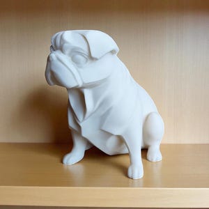 May include: A white ceramic pug dog figurine, sitting on a wooden shelf. The pug is sculpted with geometric facets, giving it a modern look. The figurine is a solid, off-white color and is set against a neutral background.