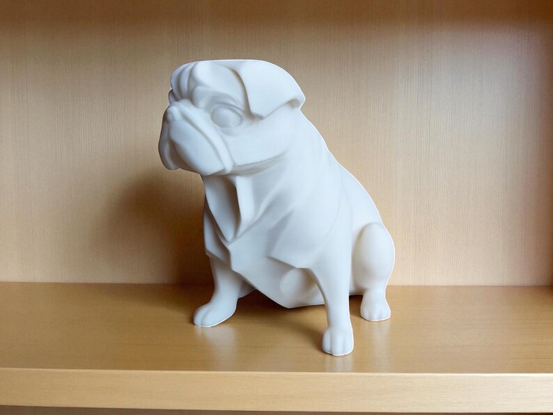 May include: A white ceramic pug dog figurine, sitting on a wooden shelf. The pug is sculpted with geometric facets, giving it a modern look. The figurine is a solid, off-white color and is set against a neutral background.