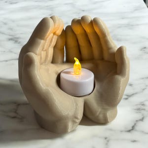 Puede incluir: Un portavelas beige en forma de manos sostiene una peque&ntilde;a vela LED blanca con una llama amarilla parpadeante. El portavelas est&aacute; dise&ntilde;ado para parecer dos manos juntas. El fondo es una superficie con un patr&oacute;n de m&aacute;rmol blanco y gris.