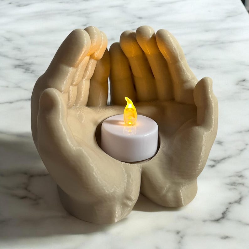 Puede incluir: Un portavelas beige en forma de manos sostiene una peque&ntilde;a vela LED blanca con una llama amarilla parpadeante. El portavelas est&aacute; dise&ntilde;ado para parecer dos manos juntas. El fondo es una superficie con un patr&oacute;n de m&aacute;rmol blanco y gris.