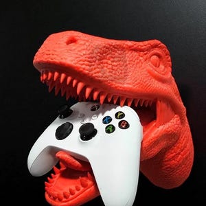 May include: A bright red dinosaur head-shaped game controller holder. The dinosaur's mouth is open, holding a white game controller with black buttons and colored action buttons. The background is black.