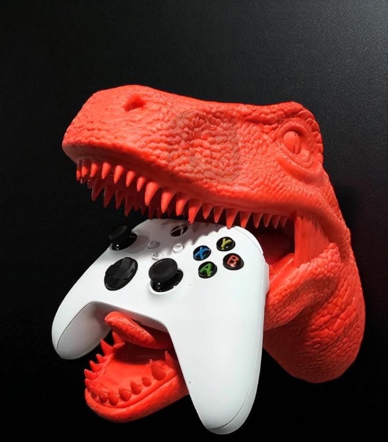 May include: A bright red dinosaur head-shaped game controller holder. The dinosaur's mouth is open, holding a white game controller with black buttons and colored action buttons. The background is black.