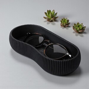 May include: A black, oval-shaped tray with a ribbed exterior holds a pair of sunglasses. The sunglasses have dark lenses and a brown and silver frame. Three small succulents are arranged in the background.