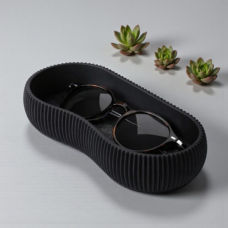May include: A black, oval-shaped tray with a ribbed exterior holds a pair of sunglasses. The sunglasses have dark lenses and a brown and silver frame. Three small succulents are arranged in the background.