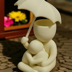 May include: A cream-colored figurine depicting a parent figure holding a child, both sheltered under a small umbrella. The figures are seated, with the parent embracing the child. The background shows a window with flowers.