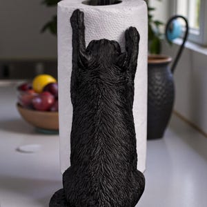 May include: A black cat-shaped paper towel holder. The cat is sculpted in a climbing pose, with its paws reaching up to hold the paper towel roll. A black candle-shaped finial with a yellow flame sits atop the roll. The holder is on a white countertop.