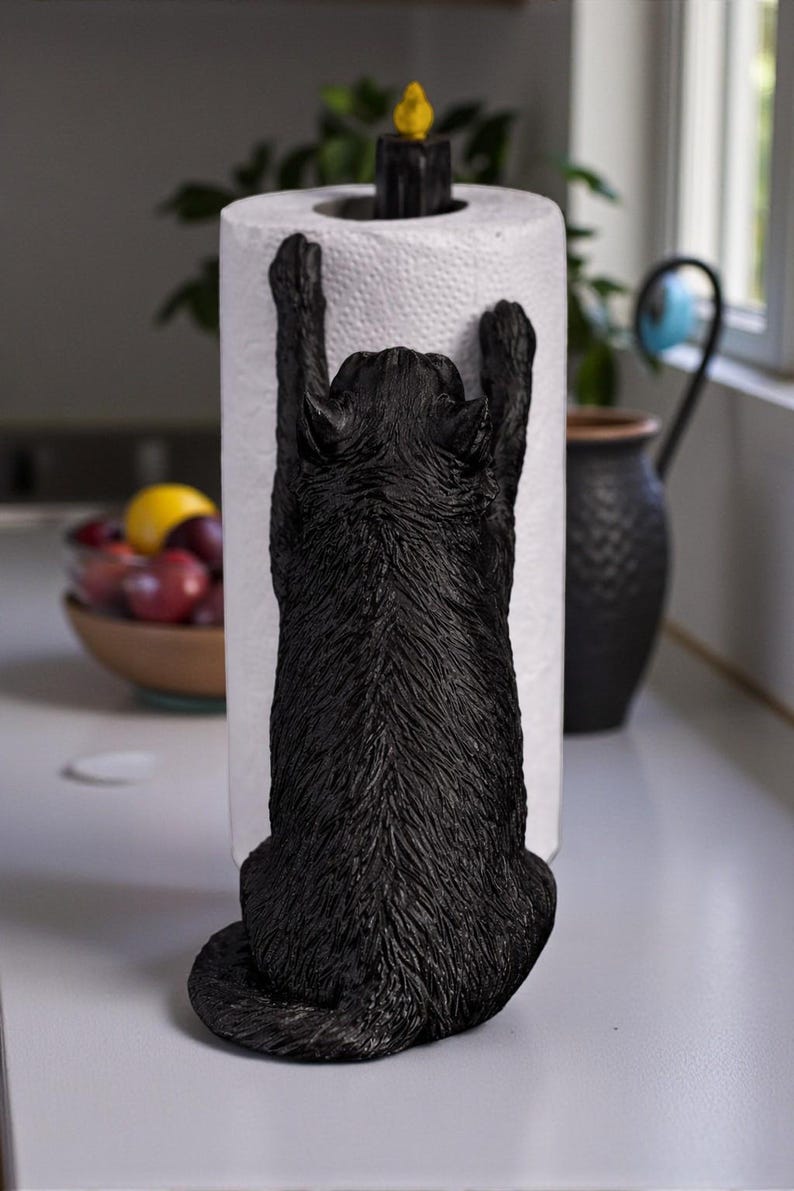 May include: A black cat-shaped paper towel holder. The cat is sculpted in a climbing pose, with its paws reaching up to hold the paper towel roll. A black candle-shaped finial with a yellow flame sits atop the roll. The holder is on a white countertop.