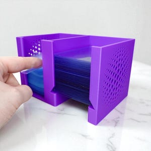 May include: A vibrant purple card holder with a geometric design on the side. The holder is divided into sections and holds a stack of blue cards. A hand is shown interacting with the cards.