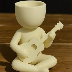 Puede incluir: Figura de color crema de una persona sentada con las piernas cruzadas, tocando una pequeña guitarra. La figura tiene una cabeza grande y abierta, diseñada para sostener una planta. La guitarra tiene un agujero de sonido redondo. La figura es de material liso y sólido.