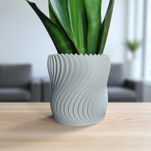 May include: A gray, wave-patterned planter holding a green plant with long, upright leaves. The planter sits on a light-colored wooden surface. The design of the planter is modern and decorative.