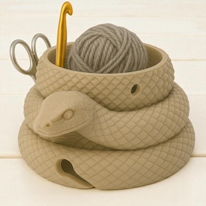 May include: A beige snake-shaped yarn bowl with a ball of gray yarn, a gold crochet hook, and silver scissors. The bowl has a textured, scale-like pattern and a hole for the yarn. A unique and functional craft accessory.