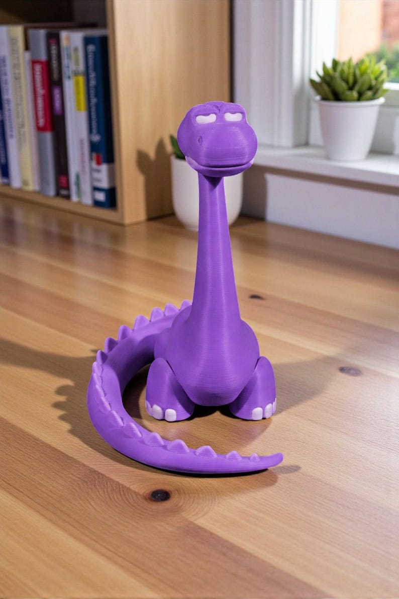 May include: A purple dinosaur figurine with a long neck and tail, and white accents on its eyes and feet. The figurine is made of a 3D-printed material and sits on a wooden surface. The dinosaur has a friendly, cartoonish appearance.