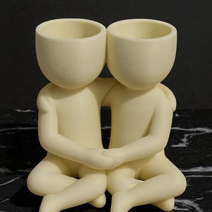 May include: A cream-coloured ceramic sculpture of two figures embracing. Each figure has a rounded head with a hollowed-out top, suggesting a planter or vase. The figures are seated with their legs crossed, arms around each other.