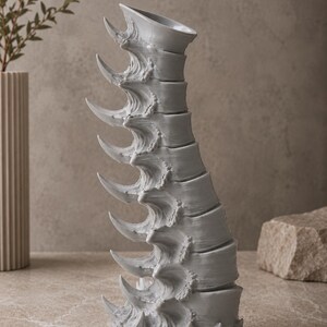 May include: A gray, sculptural vase with a unique, spine-like design. The vase features a series of curved, pointed elements along its length, creating a textured and artistic appearance. The vase is displayed on a neutral surface.