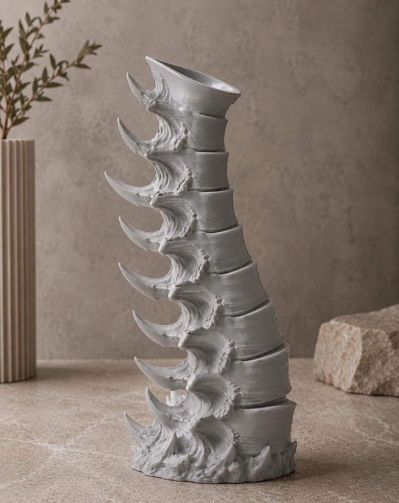 May include: A gray, sculptural vase with a unique, spine-like design. The vase features a series of curved, pointed elements along its length, creating a textured and artistic appearance. The vase is displayed on a neutral surface.