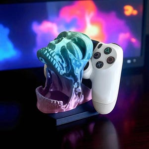 May include: A 3D-printed skull-shaped gaming controller holder. The skull is gradient-colored, transitioning from blue to purple. A white gaming controller is placed in the holder. The background is a blurred display with colorful lights.