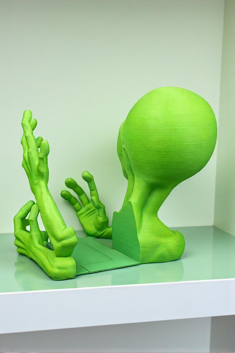 May include: A green alien-themed bookend set. The bookend features a stylized alien head and hands, all in a vibrant green color. The design is modern and unique, perfect for adding a touch of whimsy to any bookshelf.