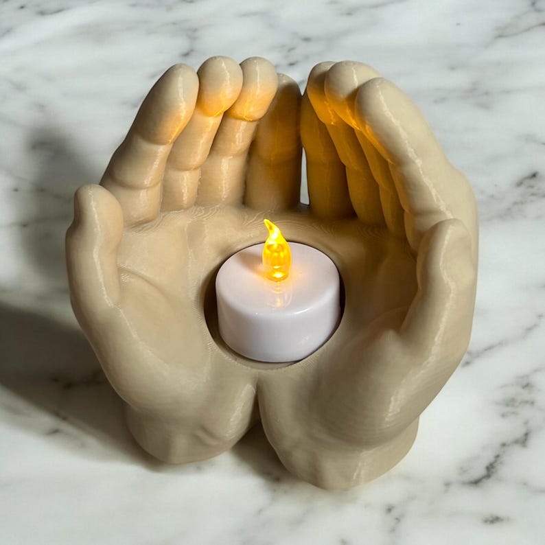 Puede incluir: Un portavelas beige con forma de dos manos que sostienen una vela blanca con una llama amarilla parpadeante. El portavelas est&aacute; hecho de un material liso y se encuentra sobre una superficie de m&aacute;rmol.