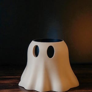 May include: A cream-colored ghost-shaped decorative vase with two oval eye cutouts and a black interior. The vase is made of a smooth material and sits on a dark wooden surface. The background is a dark gradient.