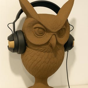 May include: A brown owl sculpture wearing black headphones. The owl has large, detailed eyes and textured feathers. The headphones have black earcups with wooden accents and a black headband. A black cord extends from the headphones.