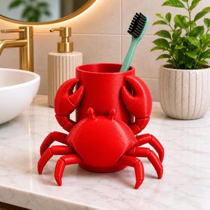 May include: A bright red crab-shaped toothbrush holder with a cylindrical cup. The holder has a detailed crab design with claws and legs. A green toothbrush with black bristles is inside the cup. The holder sits on a marble countertop.