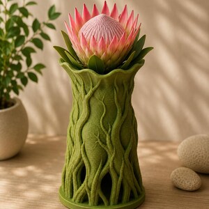 May include: A green vase with a textured, tree-like design holds a large protea flower. The flower has pink and white petals and green leaves. The vase is on a wooden surface, with a plant and decorative stones in the background.