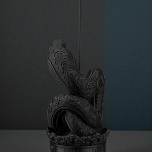 May include: A black incense holder shaped like a coiled snake, with intricate scale and pattern details. A thin incense stick rises from the center, emitting a wisp of smoke. The holder sits on a decorative base, creating a serene and artistic display.