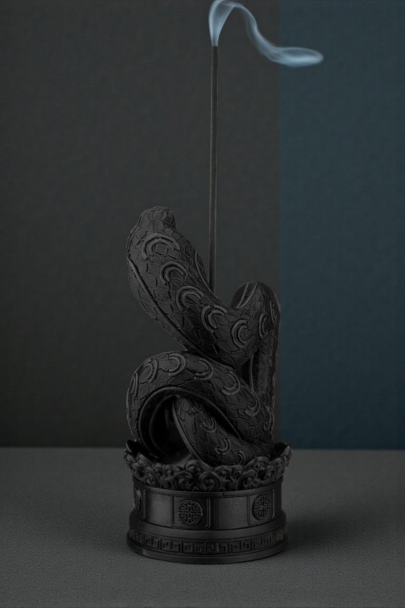 May include: A black incense holder shaped like a coiled snake, with intricate scale and pattern details. A thin incense stick rises from the center, emitting a wisp of smoke. The holder sits on a decorative base, creating a serene and artistic display.