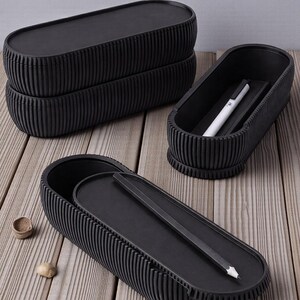 May include: Black, oval-shaped storage boxes with a ribbed design and lids. One box is open, showing a pencil and a pen. Two boxes are stacked, and another is open with a pencil inside. The boxes are on a wooden surface.
